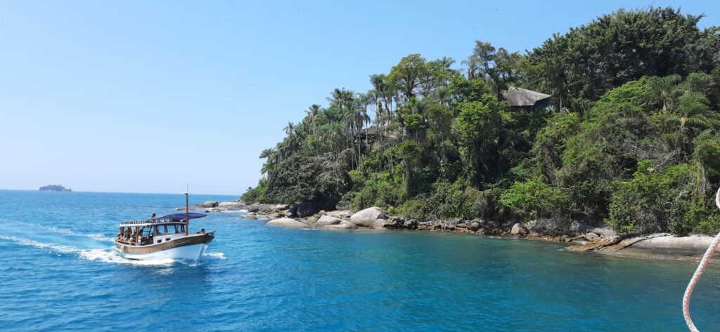 Ilha do Crepúsculo em Paraty. Do lado esquerdo um barco e o mar, do lado direito a ilha, com muitas árvores e pedras.