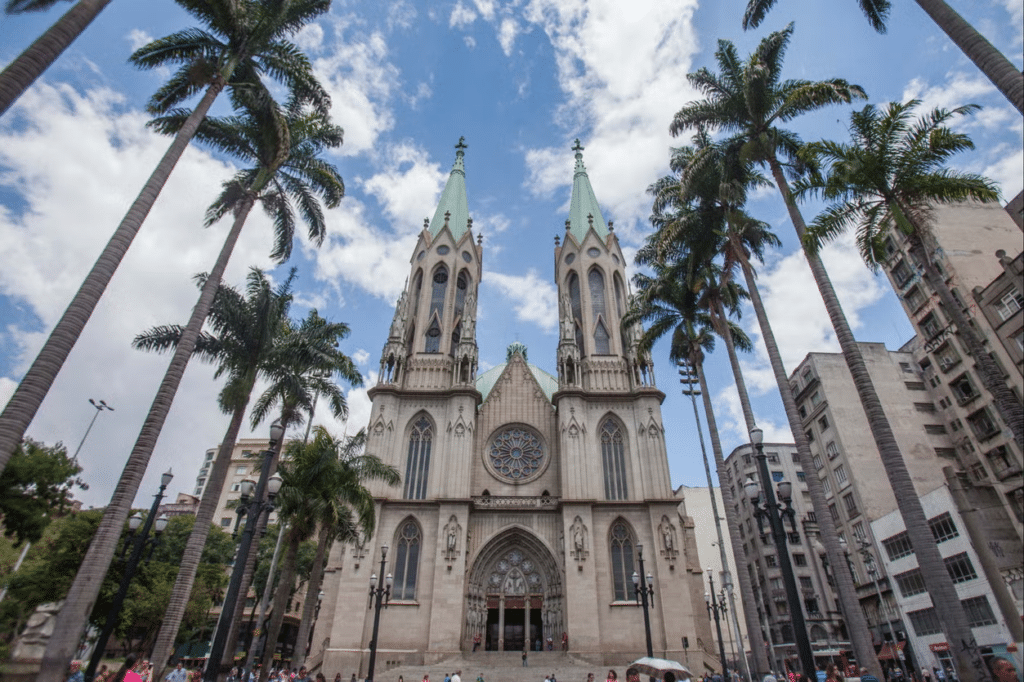 vista de frente da Catedral da Sé, em São Paulo, com estilo neogótico, vitrais imponentes e duas torres pontudas em detalhes mais claros, há palmeiras em ambos os lados e o céu está azul com nuvens