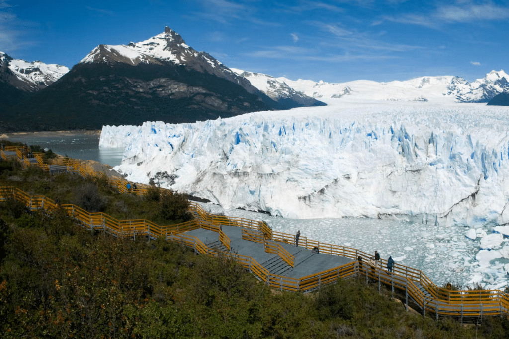 Vista panorâmica das geleiras de Perito Moreno, em El Calafate, com montanhas nevadas ao fundo, lago ao redor e uma vegetação do outro lado, há uma passarela larga cimentada com corrimões de onde é possível observar a paisagem