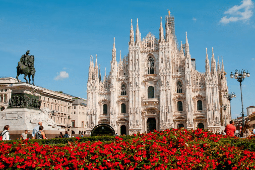 Vista do Duomo de Milão com muitas flores vermelhas à frente em um jardim, há uma escultura de um cavaleiro que aponto para a imponente catedral em estilo neogótico e fachada branca, com muitas torres pontudas e ricamente trabalhadas