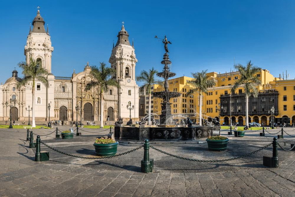Vista da Catedral na Plaza Mayor em Lima, sendo um edifício imponente e ricamente trabalhado na sua fachada, há ainda uma escultura de um anjo tocando trombeta em uma fonte alta à frente