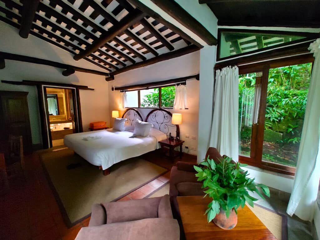 quarto do Inkaterra Machu Picchu Pueblo Hotel com uma cama de casal ao centro, uma área de estar com duas poltronas no canto direito inferior, ao lado de uma ampla janela com vista para a vegetação. Na outra ponta do quarto é possível enxergar a porta aberta para o banheiro da suíte