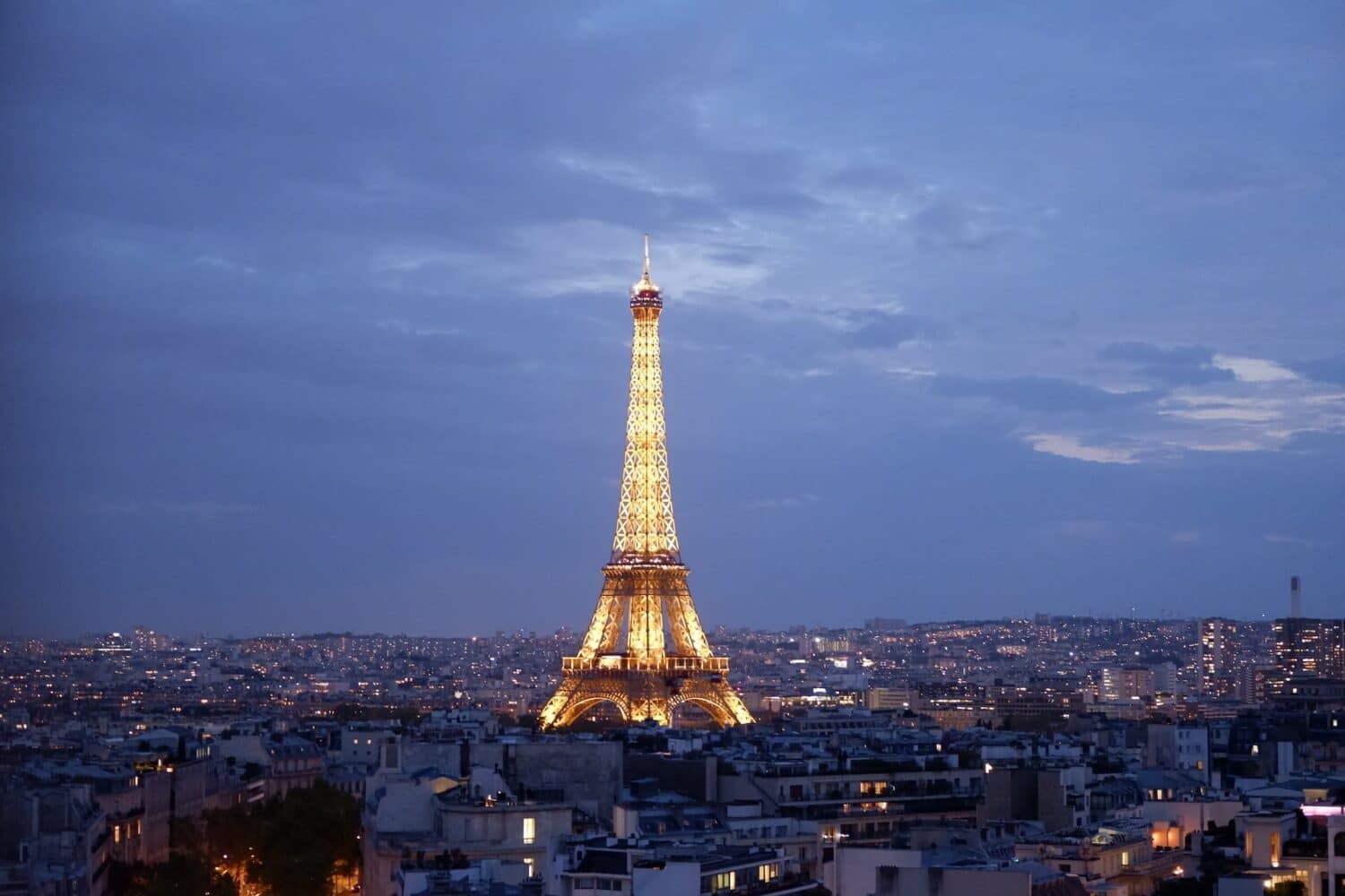 Torre Eiffel, Paris, no centro da imagem ao longe. Ela está toda iluminada durante o anoitecer com várias casas e prédios ao seu redor. - Foto: Kateryna T via Unsplash