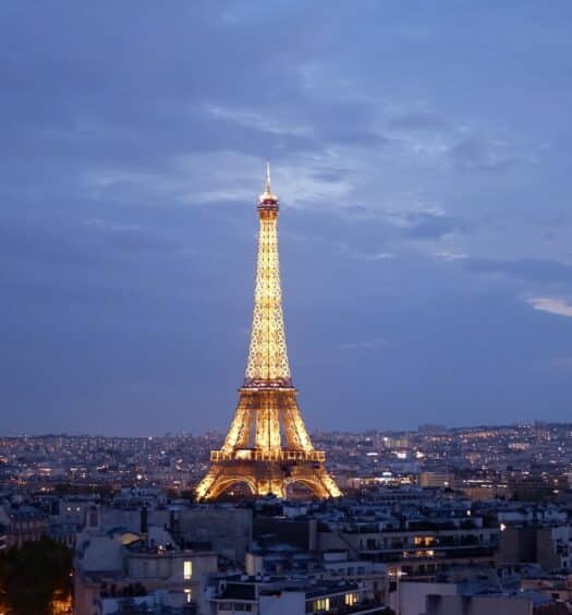 Torre Eiffel, Paris, no centro da imagem ao longe. Ela está toda iluminada durante o anoitecer com várias casas e prédios ao seu redor. - Foto: Kateryna T via Unsplash