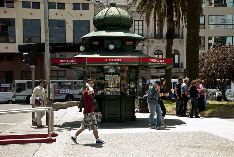 Praça em Montevidéu com um Kiosko ao meio e pessoas andando em volta. O lugar é como se fosse uma banca no meio da praça e vende chip celular Uruguai