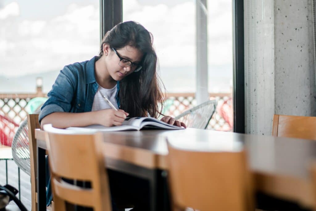 Uma mulher sentada em uma mesa de madeira, ela está com a cabeça baixa olhando um livro e segurando uma caneta em uma das mãos