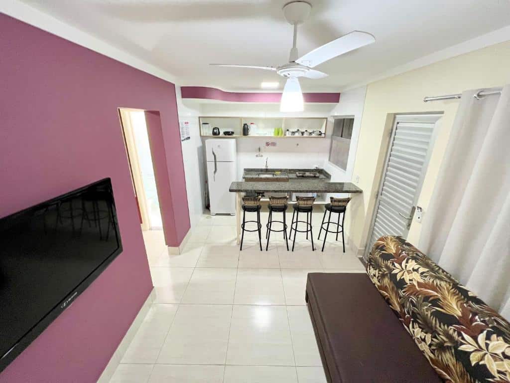 Sala de estar do L'acqua diroma I, II, III, IV e V. Do lado direito um sofá cama com uma porta e uma cortina atrás. De frente uma televisão e o corredor pro quarto. No fundo uma bancada de pedra com quatro bancos, e uma mini cozinha com pia, fogão e geladeira.