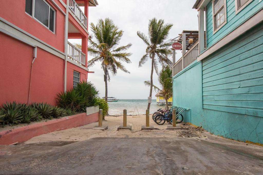 Foto tirada na rua do La Casa de Paz Hotel mostrando sua proximidade com a praia. A imagem retrata a lateral da hospedagem, e está entre um prédio vermelho à esquerda e outro azul na direita. Na frente, em poucos metros, vemos algumas bicicletas estacionadas, coqueiros e o mar.