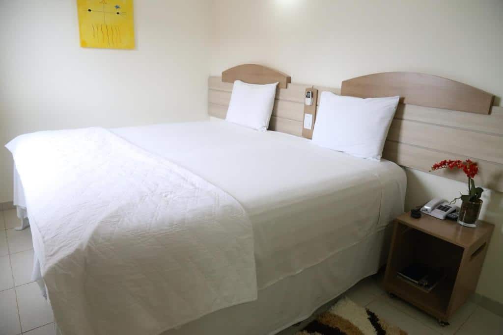 Quarto do Larison Hotéis. Uma cama de casal no meio, do lado direito uma cômoda com telefone e um vaso de flor. Foto para ilustrar post sobre hotéis em Porto Velho.