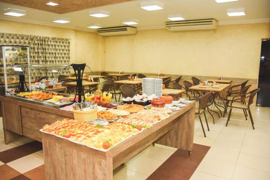 Uma ilha com café da manhã do Larison Hotéis. Uma mesa cheio de frutas, frios, pães e bolos. No fundo, mesas com cadeiras.