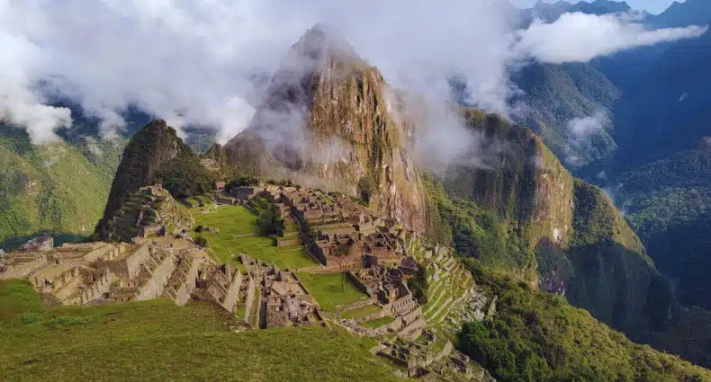 vista das montanhas de Machupicchu a uma certa distância. Há algumas nuvens cobrindo a montanha mais alta. O céu está azul e a grama bem verde.