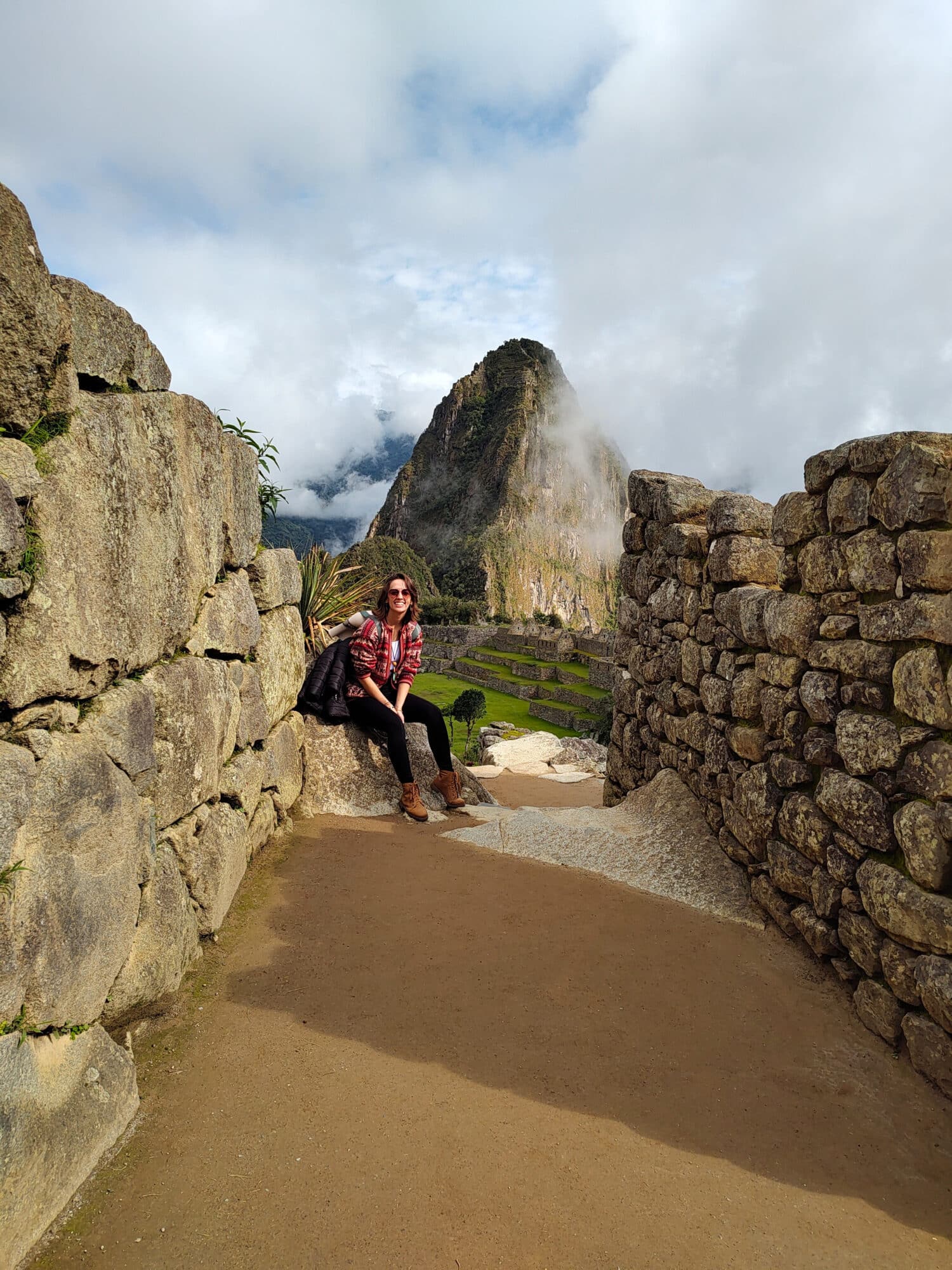 mulher sentada em cima de uma pedra no chão, usando um cardigã artesanal em tons vermelhos. Nas duas laterais é possível enxergar muros de pedras, e ao fundo do corredor, há uma parte da montanha de Machupicchu