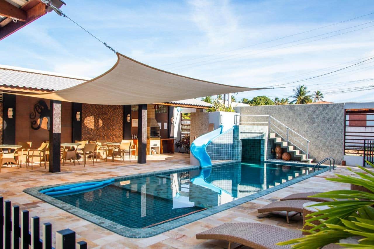 Quintal de uma casa de temporada em Maragogi, Alagoas, com uma piscina, escorregador na piscina e uma escada para dar acesso, e á esquerda tem uma área coberta com mesas e cadeiras, e uma churrasqueira