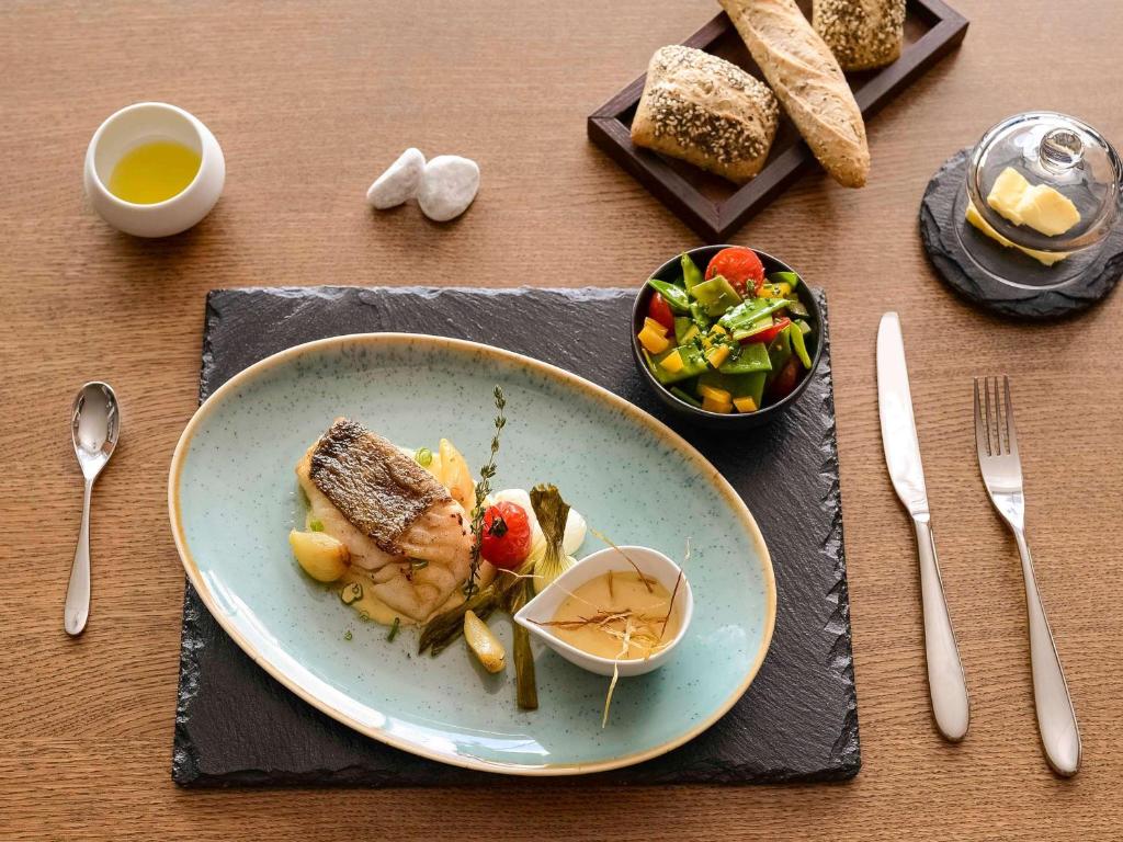 Café da manhã no Mercure Geneva Airport. A mesa é de madeira clara, possui um prato azul oval com uma refeição sofisticada e uma cumbuca preta com salada ao lado. Os talheres estão dispostos do lado do prato. Um pouco à frente, há outra min bandeja com pães e um cubo de manteiga.