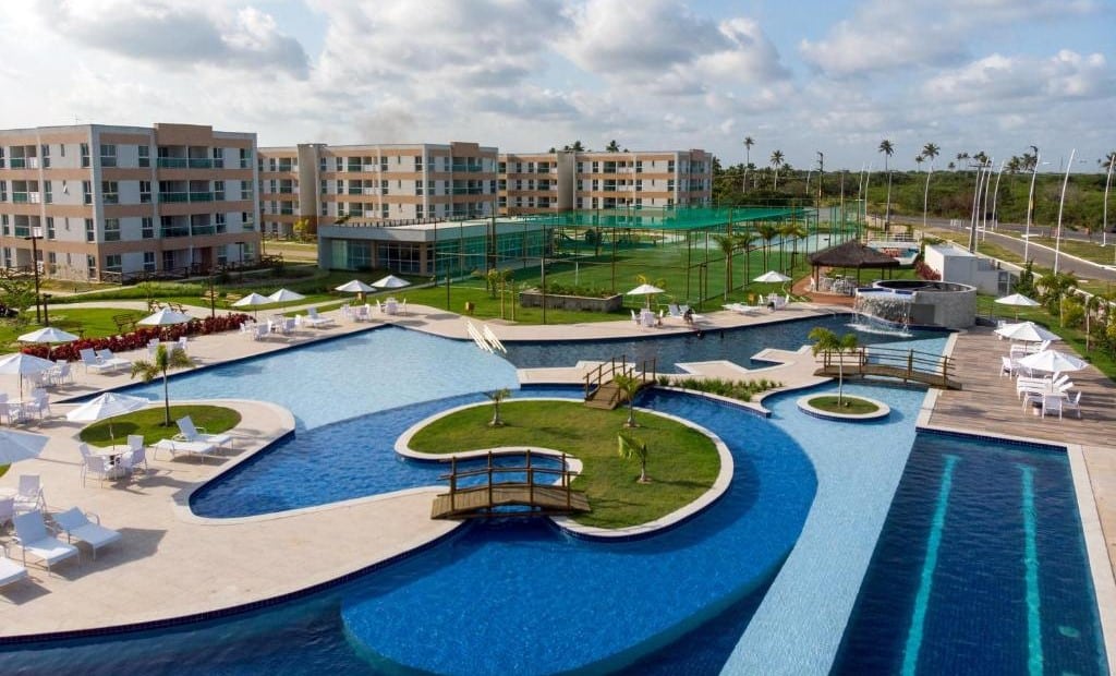 Vista da área externa do Muro Alto Condomínio Clube. Uma piscina grande no meio, com pontes, ilha de jardim, e ao redor cadeiras de tomar sol. No fundo um campo, e os apartamentos.