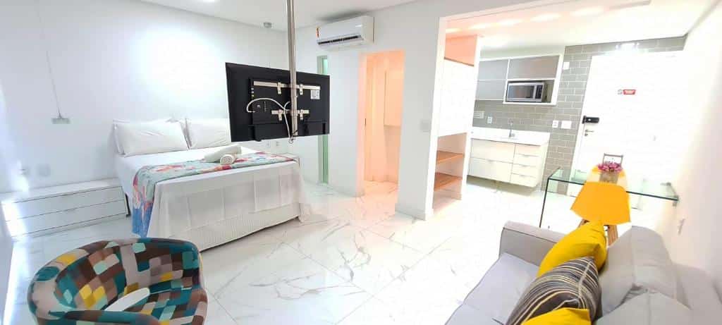 Apartamento do Muro Alto Condomínio Clube. Um sofá do lado direito, no meio uma poltrona e uma televisão, no lado esquerdo uma cômoda, uma cama de casal. No fundo, no lado esquerdo um ar-condicionado e uma porta do banheiro. No canto direito a cozinha e a porta do apê.