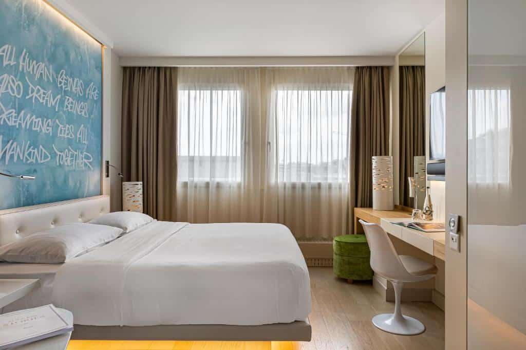 Foto de um N’vY Manotel, para ilustrar o post de hotéis em Genebra. Vemos a lateral do quarto, a cama de casal está encostada na parede esquerda, que há um quadro azul. Na parede do fundo, há uma janela. E no lado direito do quarto, de frente para a cama, há uma escrivaninha com cadeira, abajur, e um puff.