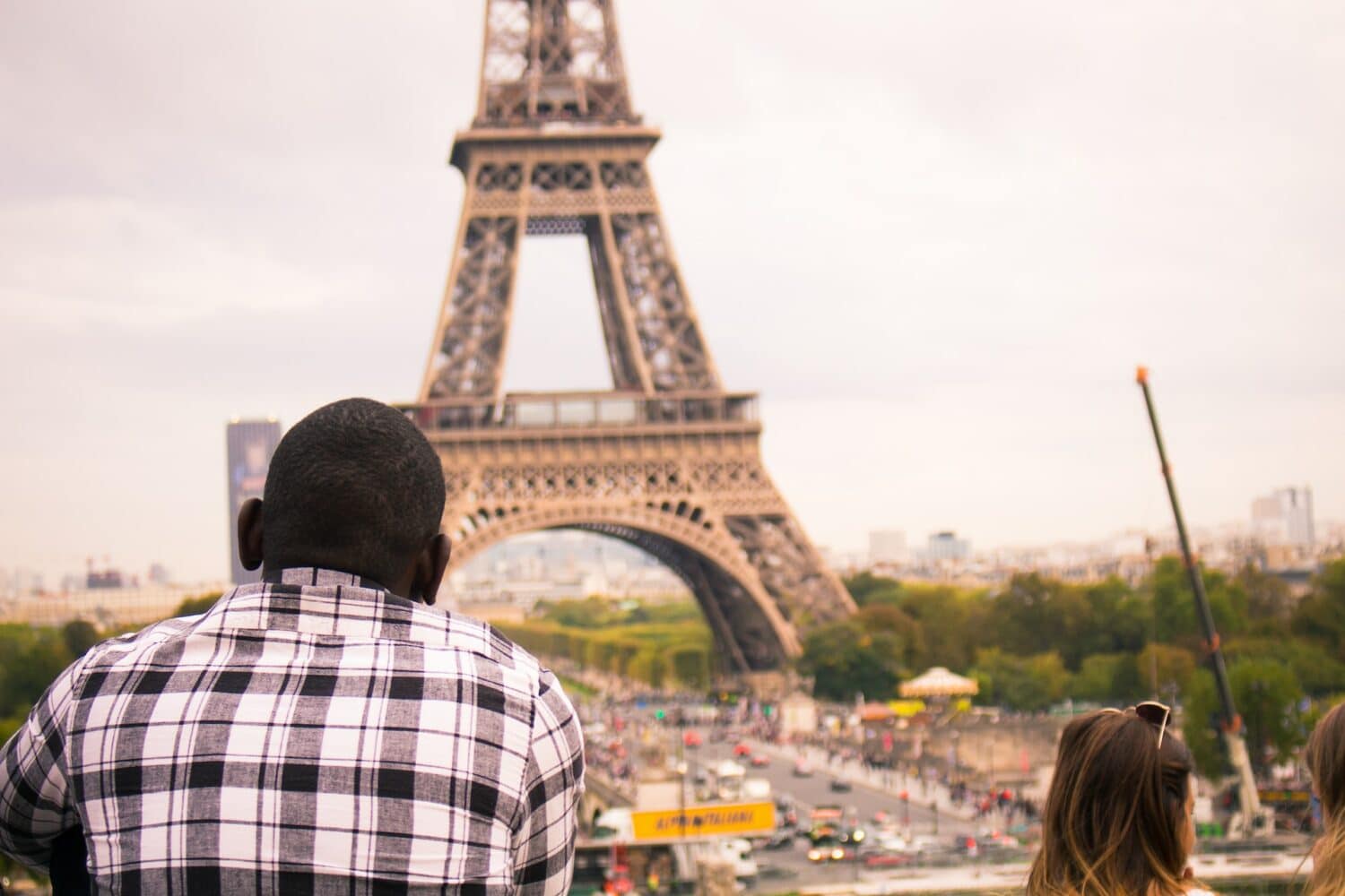 Homem negro de costas para a foto olhando a Torre Eiffel logo em frente. Ele usa uma camisa xadres e está num lugar alto. Ao seu lado, duas meninas de cabelos claros conversam entre si. É possível ver carros passando na rua abaixo. Essa foto ilustra o post sobre o melhor seguro viagem Europa. - Foto: orva studio via Unsplash