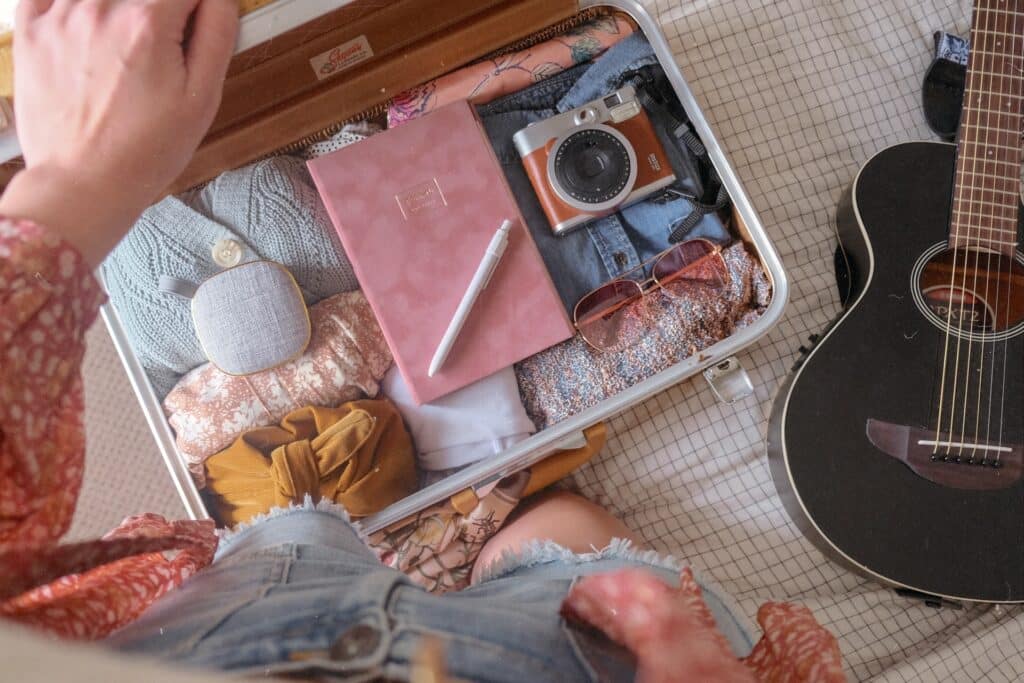 Uma mala aberta com itens de roupas, câmeras fotográficas, óculos de sol e um caderno, sob uma cama ao lado de um violão preto, para representar seguro saúde internacional