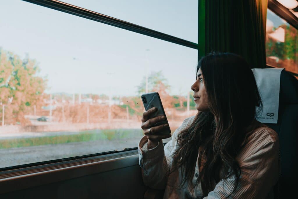 Mulher branca sentada numa poltrona de transporte público no lado direito da imagem, segurando um celular com a mão direita enquanto olha pela janela que dá vista para algumas árvores e prédios antigos em Porto, Portugal.