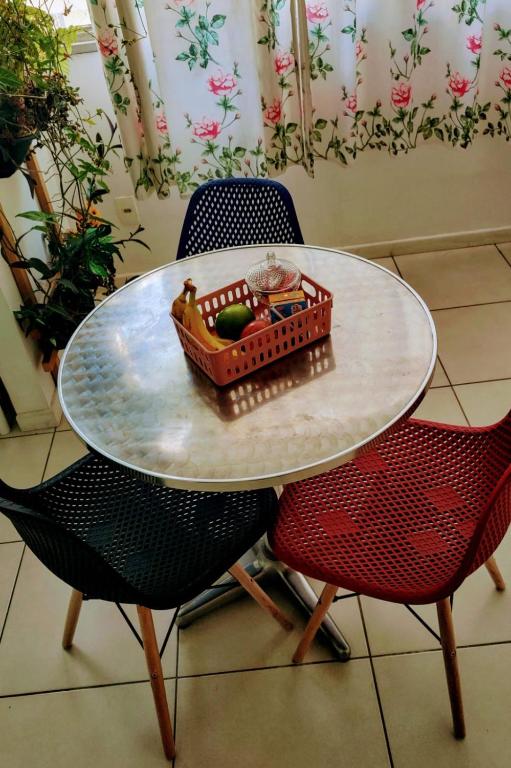 Mesa da cozinha no Pertim de Tudo. Ela é redonda e tem três cadeiras. Uma cesta com frutas está em cima dela.