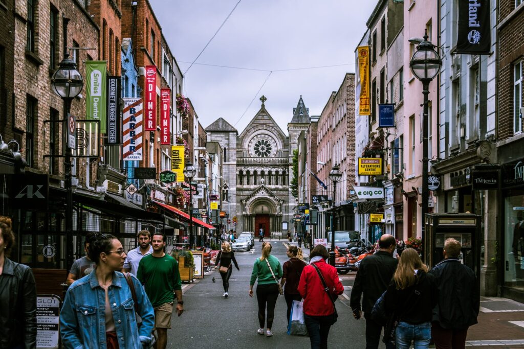 Uma rua movimentada com muitas lojas e pessoas caminhando, ao fim da rua está a St Ann's Church of Ireland em Dublin