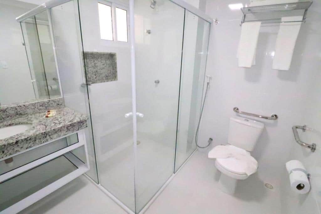 Banheiro do Piazza Acqua Park. Do lado direito um vaso sanitário com barras de apoio. No meio um box, do lado esquerdo uma pia com espelhos.