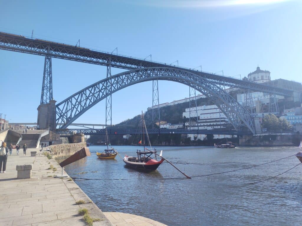 Ponte D. Luís I durante o dia com barcos do lado direito da imagem dentro do rio e ao fundo a ponte.
