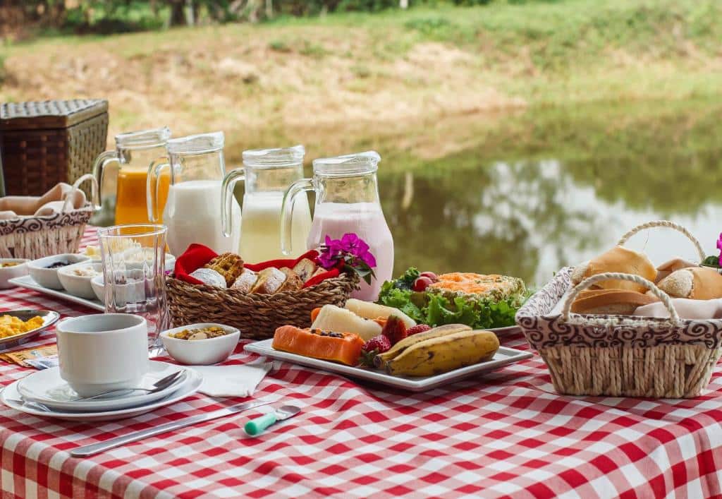 Café da manhã da Pousada Equilíbrio dos Canyons. Em cima da mesa há jarras com sucos e iogurte, também há pratos com frutas e cestos com pães. No fundo da imagem é possível ver um pedaço de um lago.