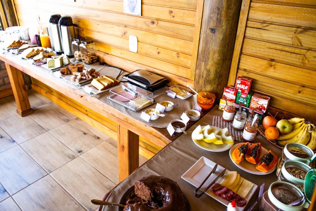 Mesas de café da manhã da Pousada Morada da Serra. As mesas estão dispostas em um formato em L e está encostada na parede. Em cima das mesas estão pratos com frutas, bolos, frios, garrafas com café e sucos.