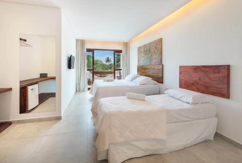 Uma cama de solteiro e uma cama de casal do lado direito da Pousada Quatro Estações. No lado esquerdo uma televisão e um espelho refletindo um balcão com frigobar. No fundo uma sacada com rede.