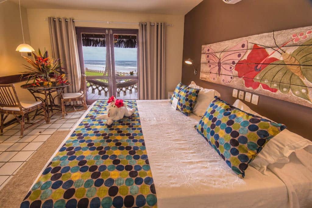 Quarto da Pousada Tabapitanga. Uma cama de casal do lado direito, do lado esquerdo uma mesa com duas cadeiras e um lustre. No fundo, a porta de vidro para a varanda com rede e vista do mar.