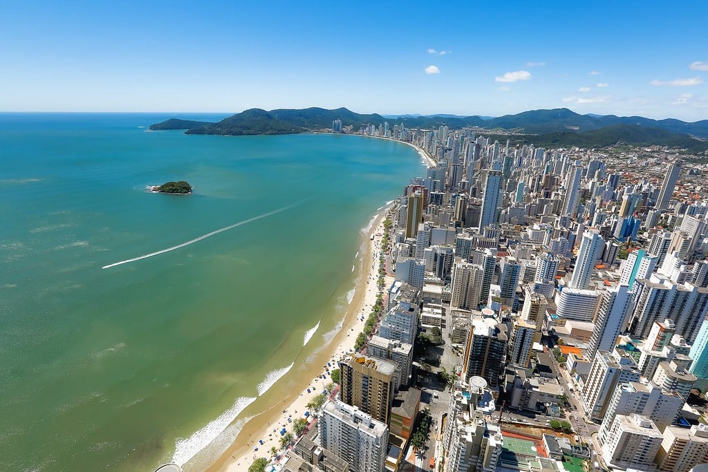 Imagem de cima da Praia Central de Balneário Camboriú. Do lado direito diversos prédios, no meio a orla da praia e no lado esquerdo o mar.