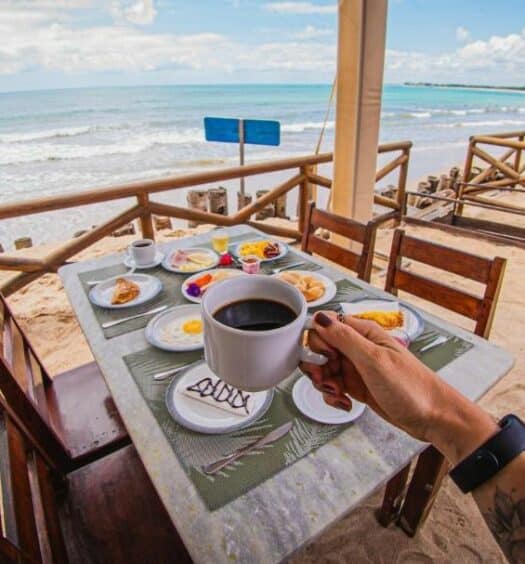 Foto do café da manhã do Privê Pontal de Maracaipe para ilustrar post sobre pousadas em Ipojuca. Uma mesa com quatro cadeiras e pratos com ovo, pães e frios. Uma mão em cima segurando uma xícara de café da manhã. No fundo, o mar.