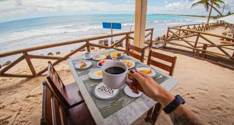 Foto do café da manhã do Privê Pontal de Maracaipe para ilustrar post sobre pousadas em Ipojuca. Uma mesa com quatro cadeiras e pratos com ovo, pães e frios. Uma mão em cima segurando uma xícara de café da manhã. No fundo, o mar.