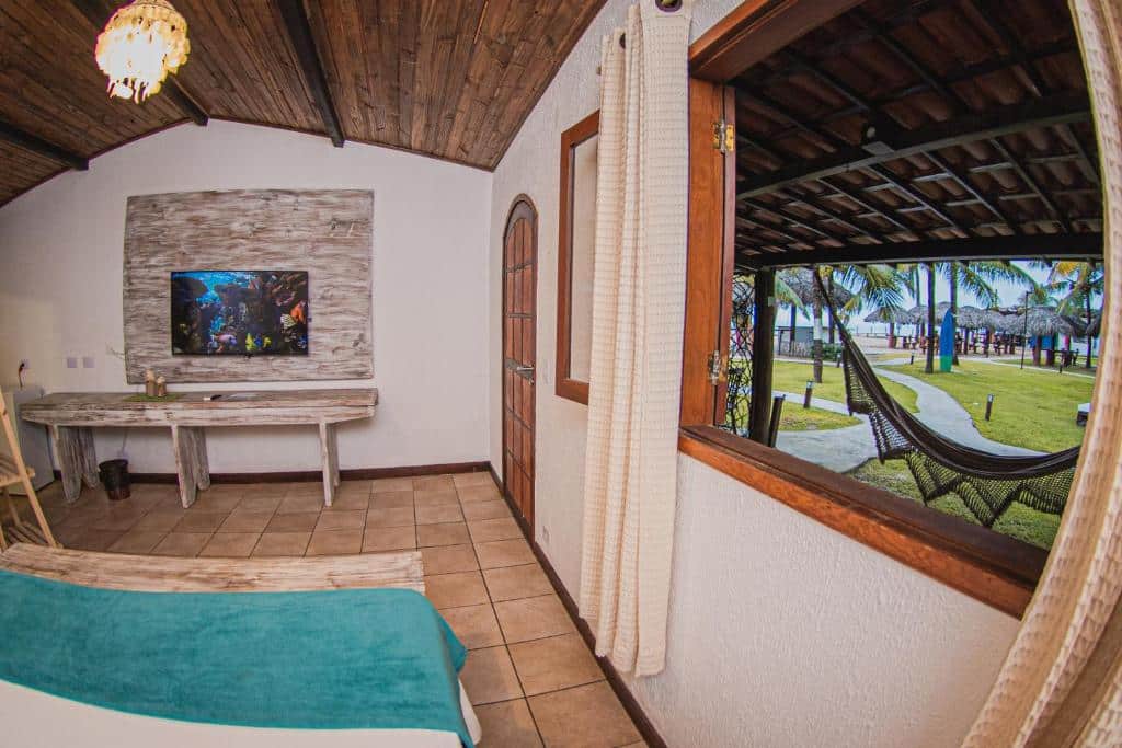 Quarto do Privê Pontal de Maracaipe. Uma cama de casal do lado esquerdo,, de frente uma mesa e uma televisão. No lado direito a janela e a porta do quarto, com uma rede na varanda e a vista do mar.