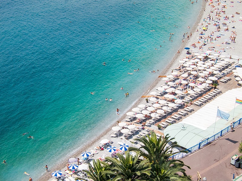 Promenade des Anglais, famosa avenida beira-mar em Nice, visto de cima. No lado esquerdo da imagem é possível ver boa parte do mar em tons de azul claro e escuro. Já no lado direito da imagem há uma faixa de pedrinhas semelhante a areia com diversos guarda-sóis e banhistas, além de um pedaço da avenida beira-mar.
