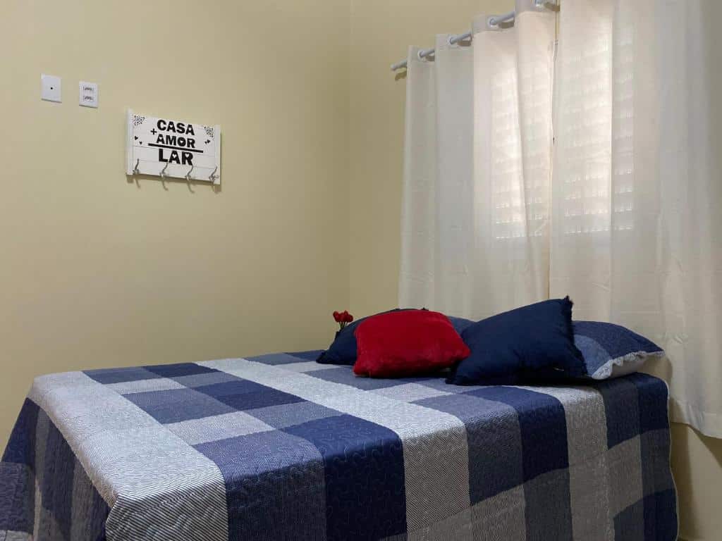 Quarto do airbnb Casa em Brotas, Refúgio e Aconchego com uma cama de casal com uma colcha e alguns travesseiros. Atrás da cama tem uma janela com cortina e na parede do lado esquerdo tem um quadro e uma tomada.