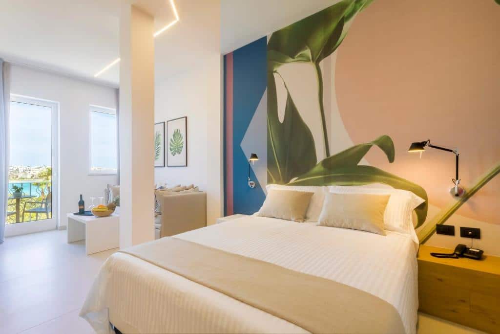 Quarto do Hotel Bellavista - Boutique Hotel com uma cama de casal, uma mesinha de madeira ao lado direito e dois abajures de parede em cada lado da cama, ao fundo do lado esquerdo tem uma pilastra, um sofá, uma mesinha com umas comidas e vinho, dois quadros na parede e uma porta de vidro que dá acesso a varanda.