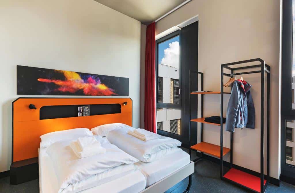 Foto de um quarto no MEININGER Hotel Genève, para ilustrar o post de hotéis em Genebra. A cama está no centro, na esquerda, e atrás há uma cabeceira laranja e um quadro com arte moderna. Na direita da cama, há uma janela e arara para roupas.