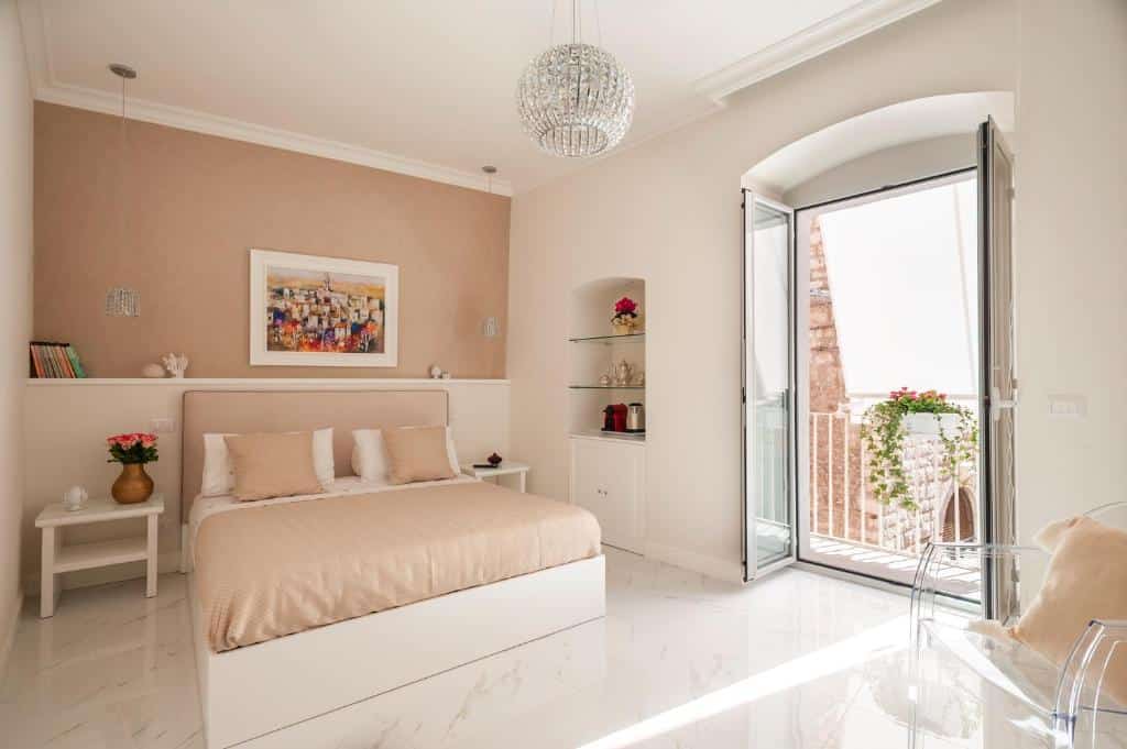 Quarto do Porta Piccola Luxury Home com uma cama de casal ao centro, do lado esquerdo tem uma mesinha com um vaso de flor e do lado direito tem algumas prateleiras com objetos. Na parede atrás da cama tem um quadros, dois lustres e alguns livros, já em frente a cama tem uma poltrona transparente com uma almofada e uma porta de vidro que dá acesso a varanda do quarto.