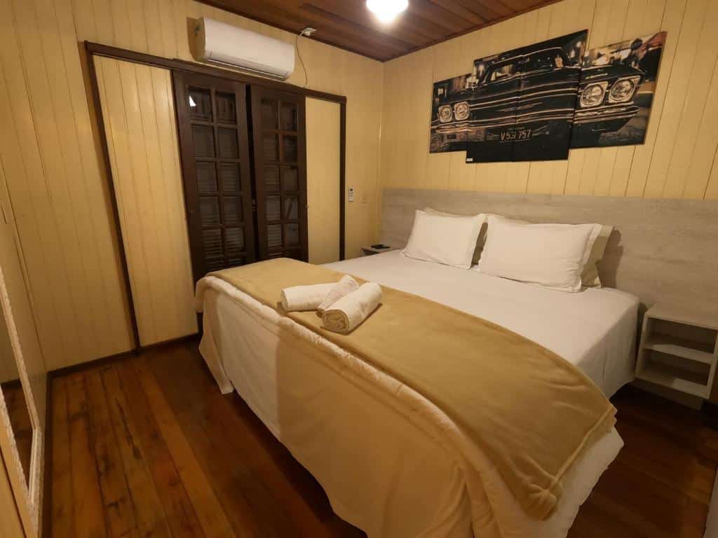 Quarto da Pousada Vó Liane. A cama está centralizada e encostada na parede do lado direito do quarto. No lado esquerdo há uma porta de madeira e um ar-condicionado em cima. Em cada lado da cama há um pequeno móvel quadrado com duas prateleiras. Imagem para ilustrar o post pousadas em Aparados da Serra.