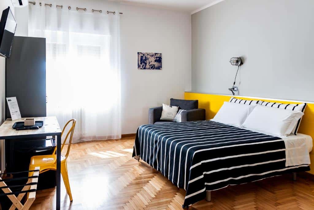 Quarto do TRAVEL STATION com uma cama de casal ao centro, do lado esquerdo da cama tem uma poltrona com almofadas, uma lâmpada na parede e um quadro do lado da janela coberta pela cortina. Já em frente a cama tem um armário, uma tv na parede e uma escrivaninha com uma cadeira.