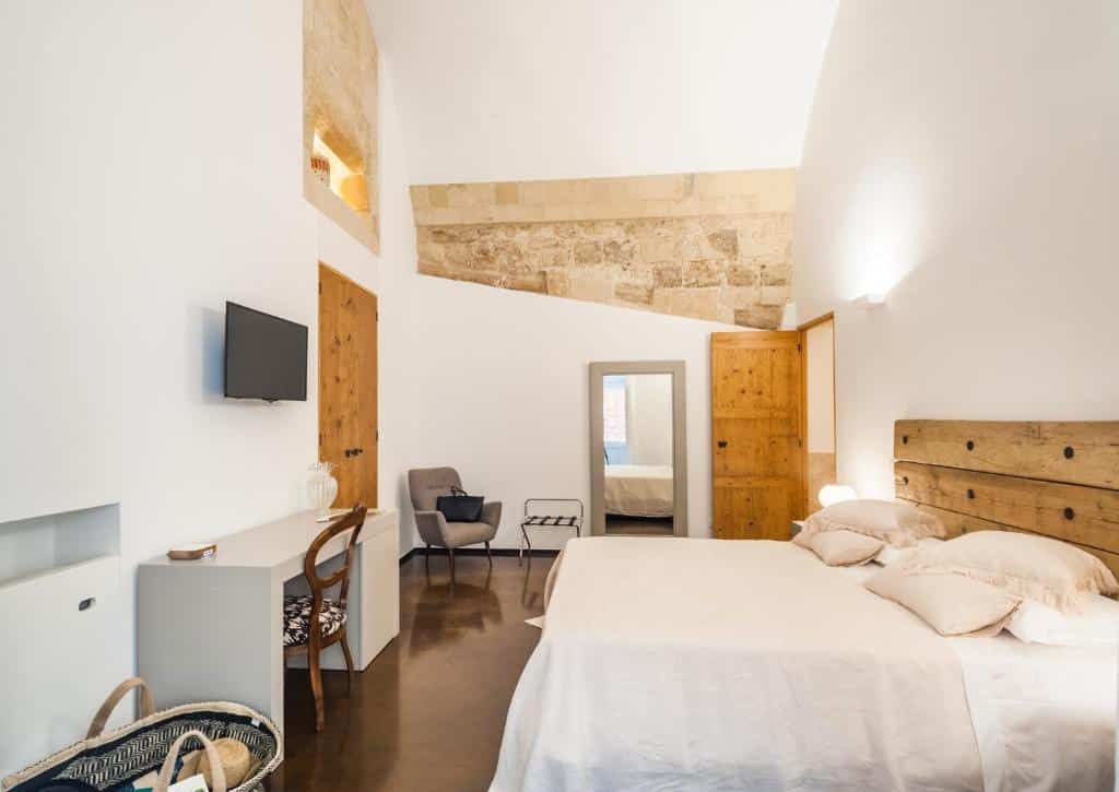 Quarto do Vico dei Bolognesi Palazzo Personè com uma cama de casal com travesseiros em cima e uma cabeceira de madeira. Em frente a cama tem uma escrivaninha com uma cadeira e uma tv em cima na parede. Ao fundo do quarto tem uma porta de madeira, uma poltrona, um espelho e outra porta de madeira aberta. As paredes do quarto são abertas brancas e tem detalhes de pedra.