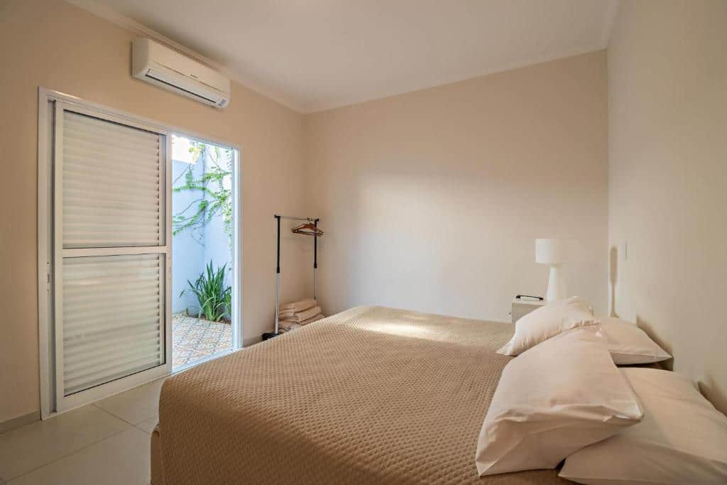 Quarto do Vila Brotas com uma cama de casal com uma colcha e alguns travesseiros em cima, do lado direito uma mesinha com um abajur e em frente a cama tem uma arara de roupa, uma porta aberta que dá acesso ao jardim e um ar-condicionado em cima da porta.