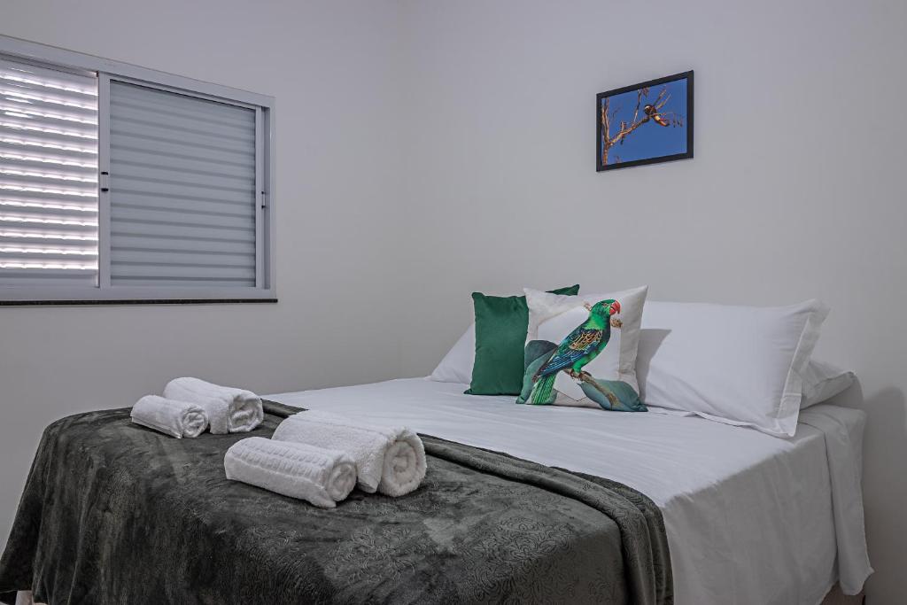 Quarto do Vila Pepira ViP com uma cama de casal com travesseiros, almofadas, edredom e algumas toalhas em cima. Na parede tem um quadro e do lado esquerdo da cama tem uma janela de alumínio branca.