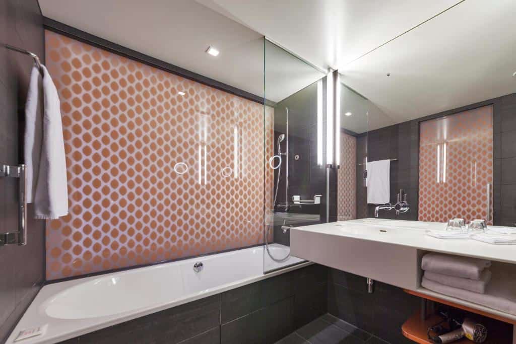 Banheiro do Radisson Blu Hotel. O ambiente é be moderno e possui azulejos cinzas. A pia está na direita, com um grande espelho, toalhas e secador de cabelo em seus compartimentos. A banheira está ao seu lado, no centro da foto. Também possui ducha inclusa.