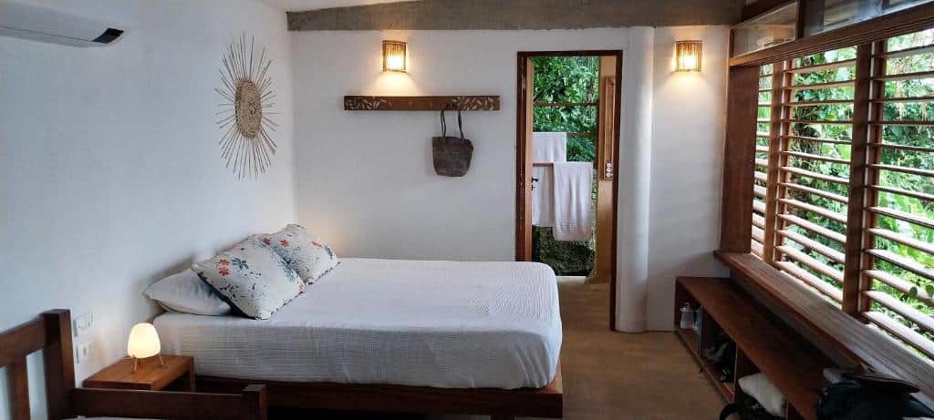 Quarto do REFUGIO na frente do mar em Ilha de Araujo. Do lado esquerdo uma cama de casal com uma mesa e uma luminária de lado. Do lado direito uma estante e a janela do quarto com vista para a praia. No fundo, a porta do banheiro. Foto para ilustrar post sobre airbnb em Paraty.