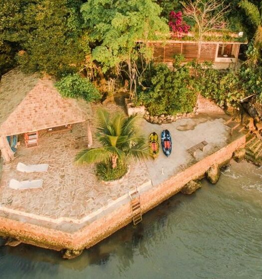 Foto da frente do REFUGIO na frente do mar em Ilha de Araujo para ilustrar post sobre airbnb em Paraty. O mar na frente, no fundo uma varanda com barquinhos, no fundo uma casa e atrás uma mata.