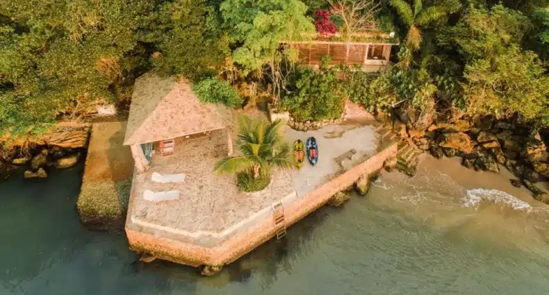 Foto da frente do REFUGIO na frente do mar em Ilha de Araujo para ilustrar post sobre airbnb em Paraty. O mar na frente, no fundo uma varanda com barquinhos, no fundo uma casa e atrás uma mata.